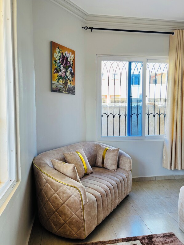 Un soggiorno angolare con un divano beige, un quadro con fiori, una finestra con una porta blu e una tenda.