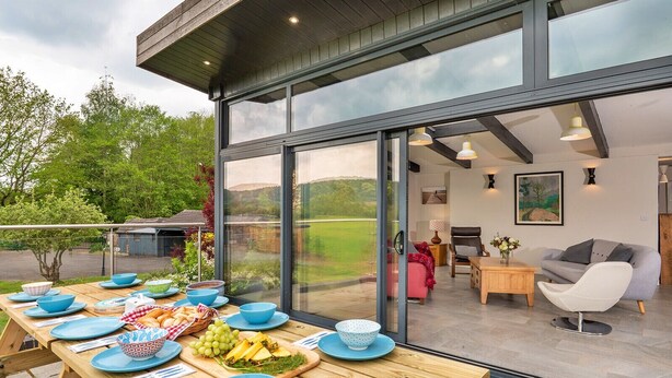 A modern outdoor dining area with a view of the countryside, a wooden table set with blue dishes, and a glass-enclosed living space with furniture. - Yonder Castle Hill (Harley)