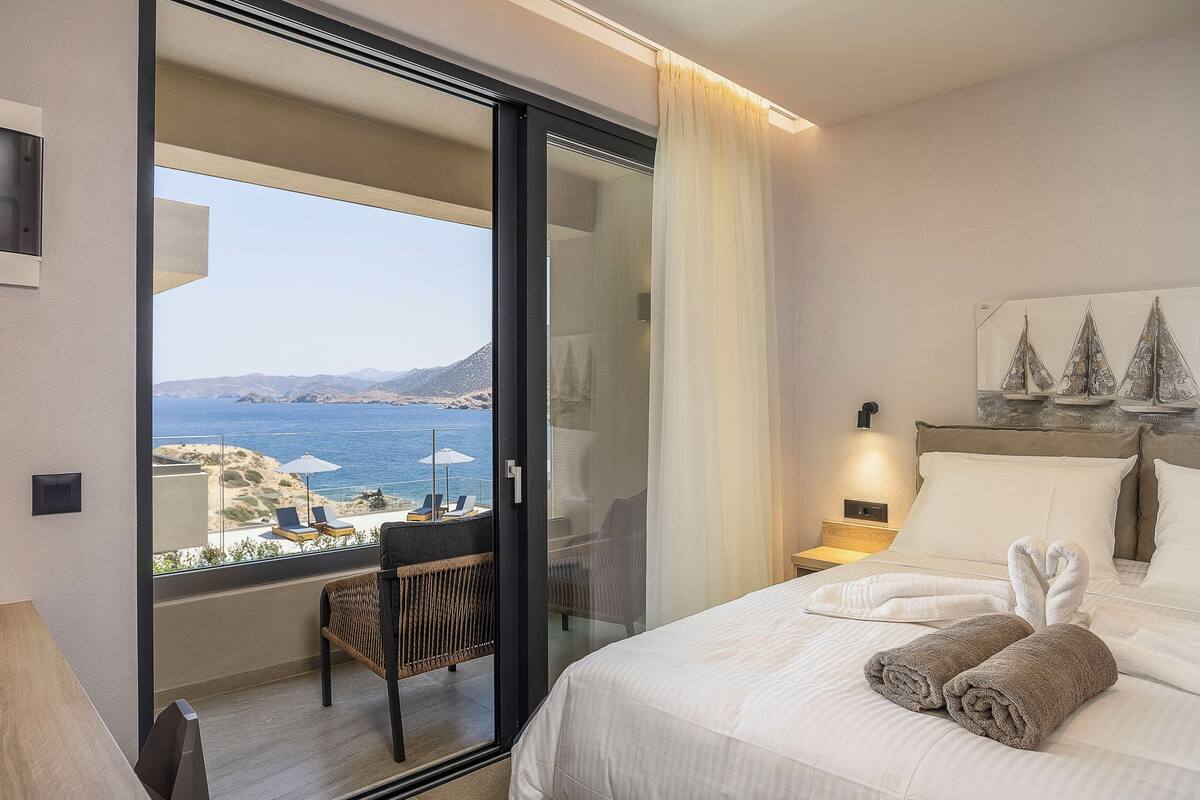 A hotel room with a bed, a chair, a nightstand, and a view of the sea and mountains.