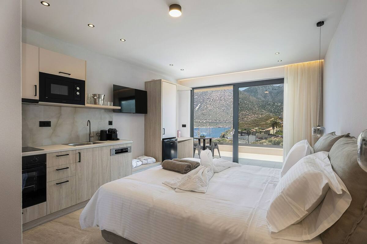A modern hotel room with a large bed, a kitchen area, and a balcony with a scenic view.