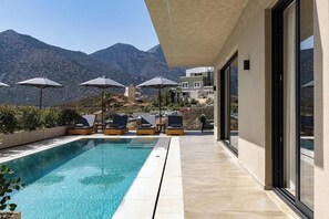 A pool area with lounge chairs and umbrellas, a clear view of mountains, and a modern building with large windows. - Celeste Bali Suites (Mylopotamos)