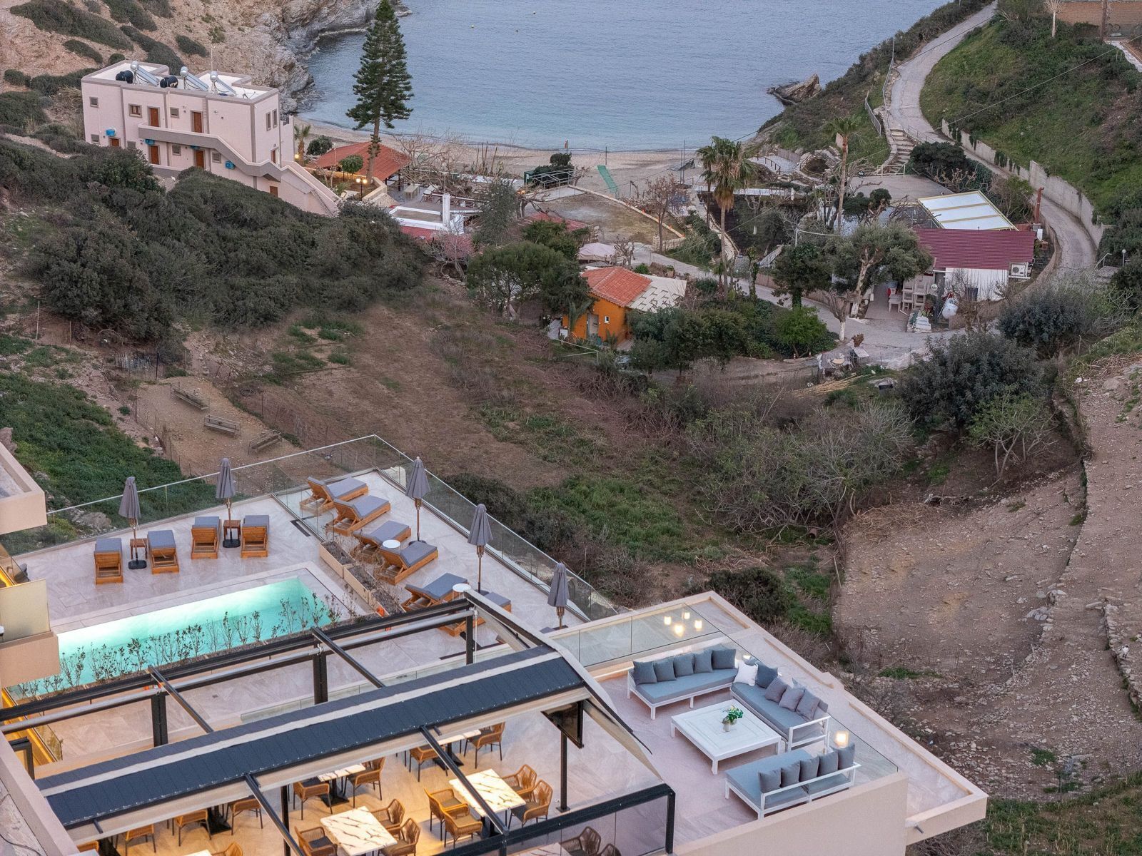 A rooftop pool area with lounge chairs and a dining area, overlooking a coastal landscape with buildings and vegetation.