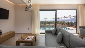 A modern living room with a large sofa, a coffee table, and a TV mounted on the wall. A large window offers a view of a pool and outdoor umbrellas. - Celeste Bali Suites (Mylopotamos)