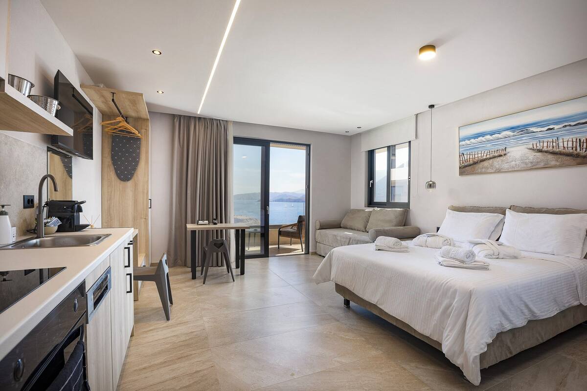 A modern hotel room with a kitchenette, a bed with towels, a seating area, and a large window with a view.
