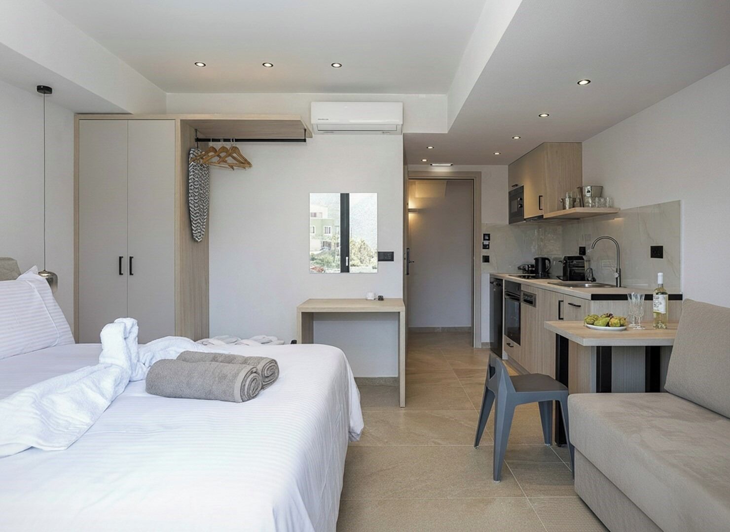 A modern hotel room with a bed, a desk, a kitchenette, and a view of the outdoors.
