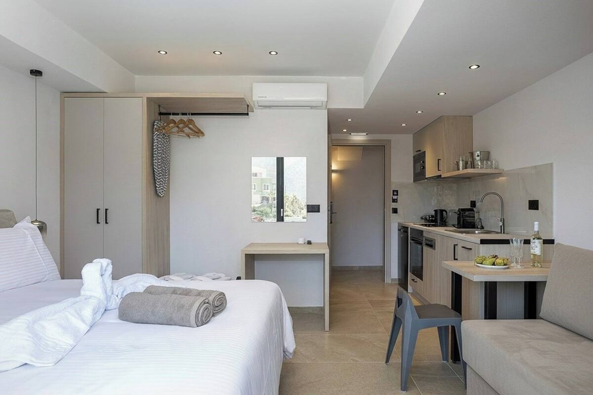 A modern hotel room with a bed, a desk, a kitchenette, and a view of the outdoors.