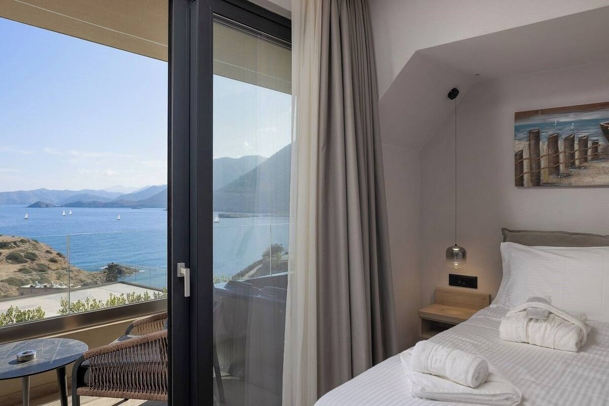 A hotel room with a large window, a bed with white linens, a small table, and a chair.