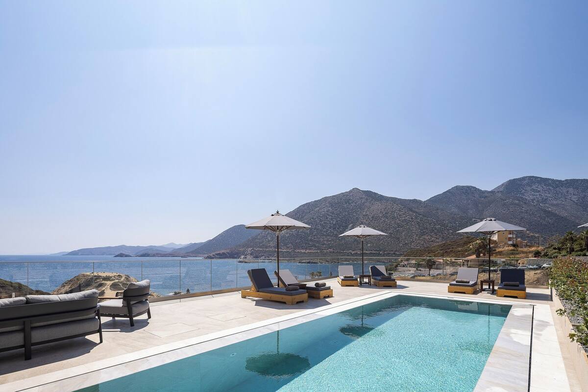 A poolside area with lounge chairs and umbrellas, overlooking a body of water and mountains.