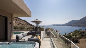 A balcony with a hot tub, seating area, and a glass railing overlooking a scenic view of the sea and hills. - Celeste Bali Suites (Mylopotamos)