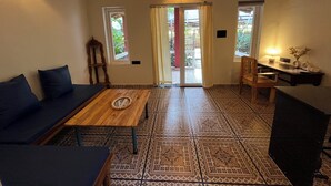 A room with a wooden coffee table, a sofa, a desk with a chair, and patterned floor tiles. - Brahma Cottages (Canacona)