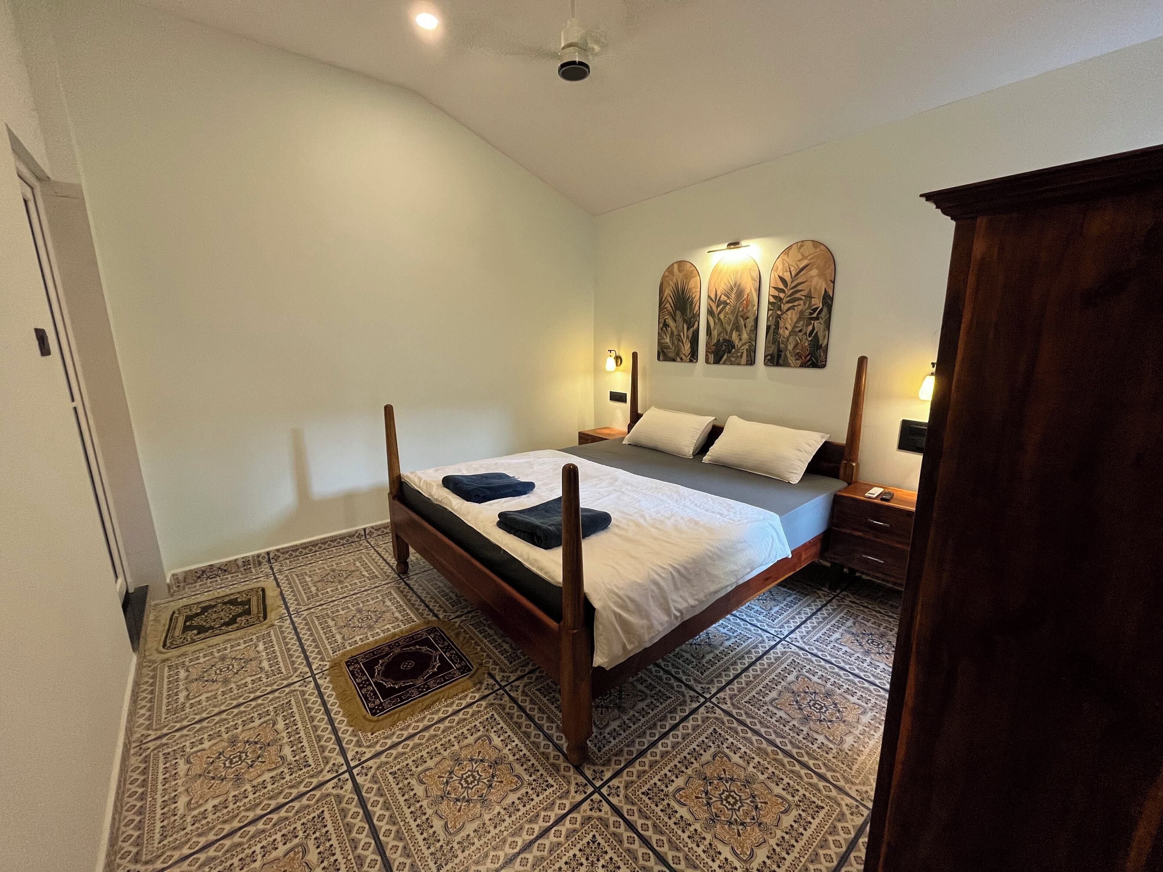 A bedroom with a wooden bed, patterned floor tiles, and a nightstand with a lamp.