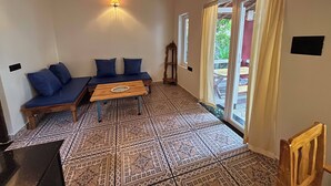 A room with a wooden ceiling, a sofa with blue cushions, a wooden table, patterned floor tiles, and a sliding glass door leading outside. - Brahma Cottages (Canacona)