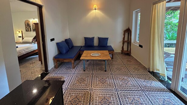 A room with a patterned floor, a wooden ceiling, a sofa with blue cushions, a wooden table, and a bed in the background. - Brahma Cottages (Canacona)