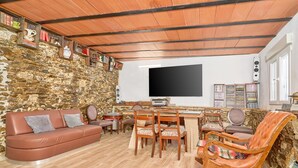 A cozy living room with a stone wall, wooden ceiling beams, a flat-screen TV, a dining table with chairs, a leather sofa, and a rocking chair. - Cortijo Palomo: Country House with Private Pool and Mountain Views (Málaga)