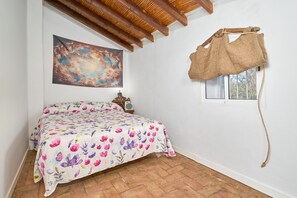 A bedroom with a bed, a painting, a rustic wall hanging, and a window. - Cortijo Palomo: Country House with Private Pool and Mountain Views (Málaga)