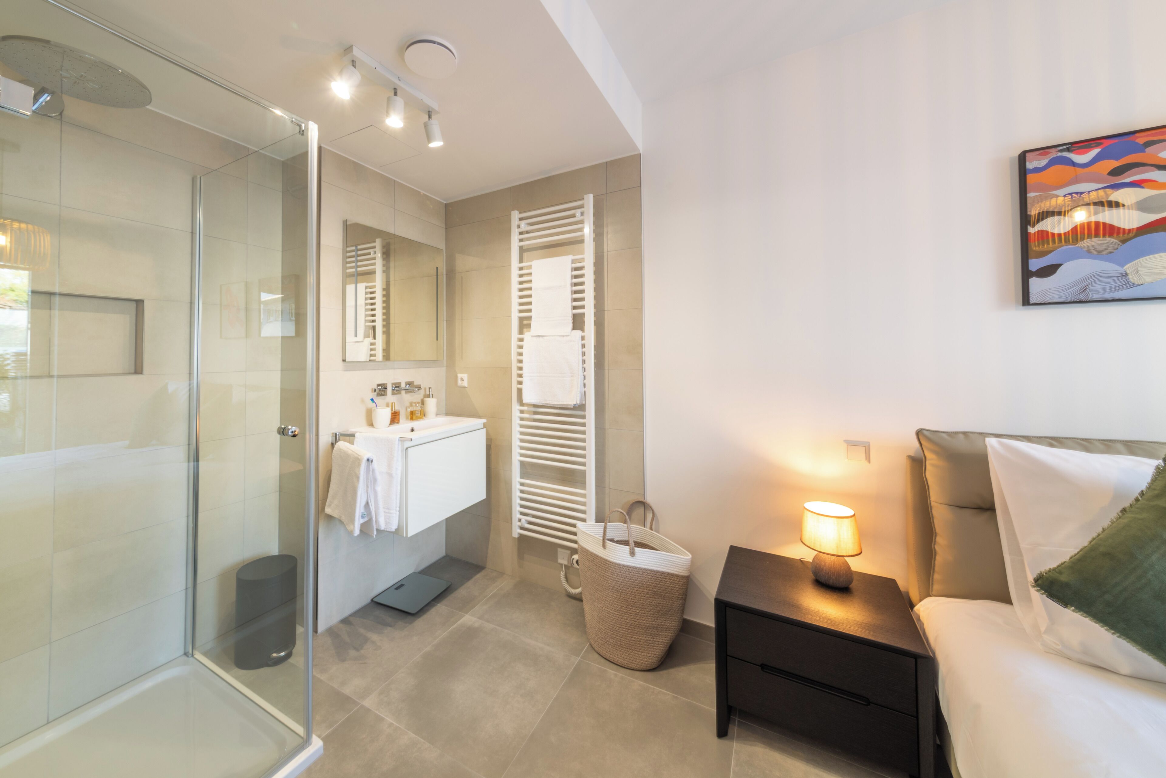Un bagno moderno con cabina doccia in vetro, un mobile lavabo bianco con specchio e un comodino con lampada.