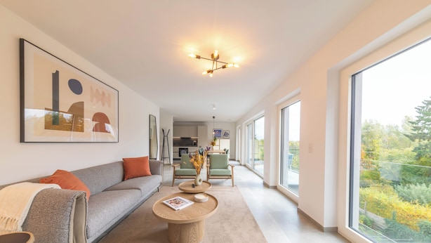 Una sala moderna con sofá, mesa de centro y sillas, ventanas amplias con vistas, y una cocina al fondo.
