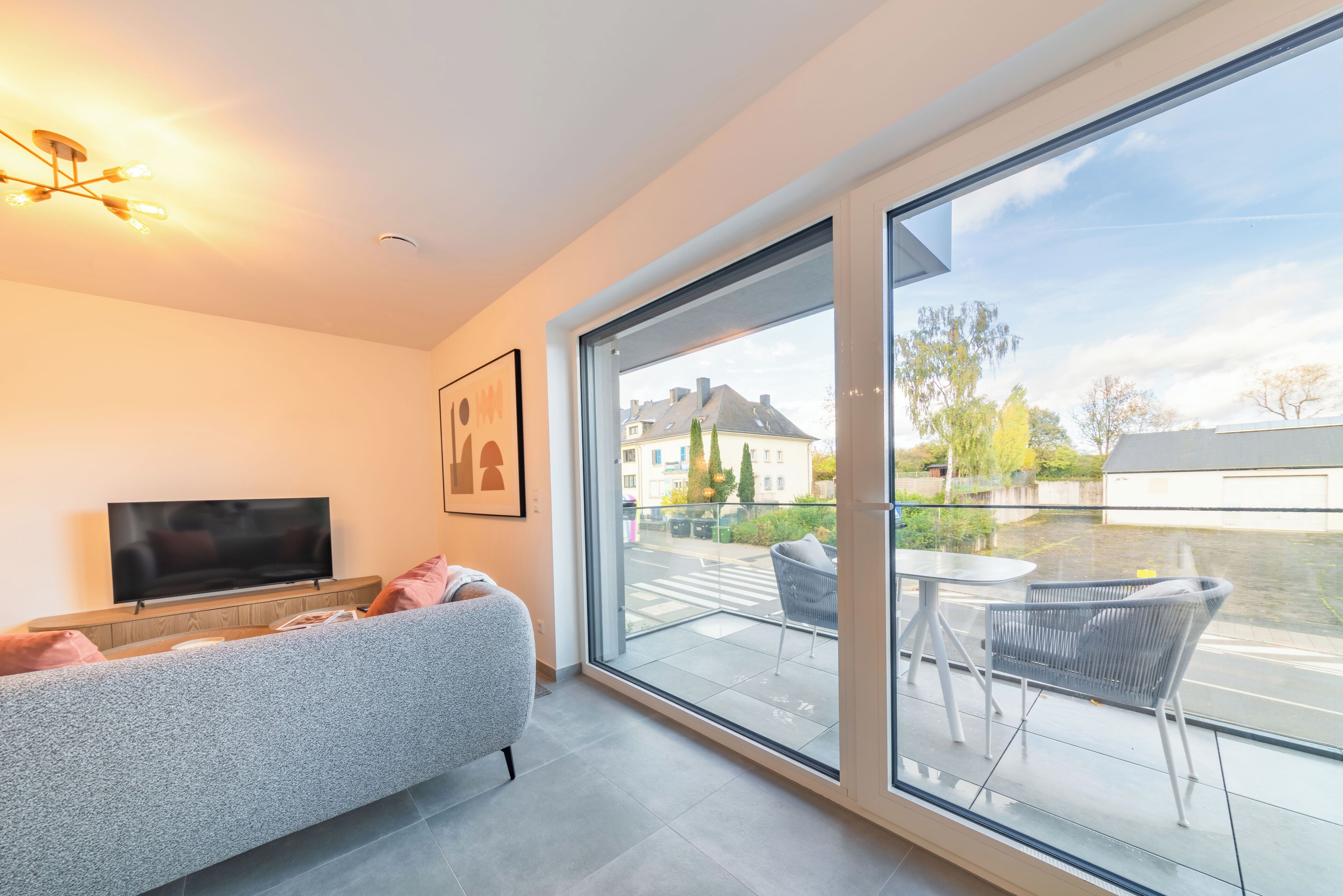 A modern living room with a sofa, a flat-screen TV, and a large sliding glass door leading to an outdoor balcony with chairs.