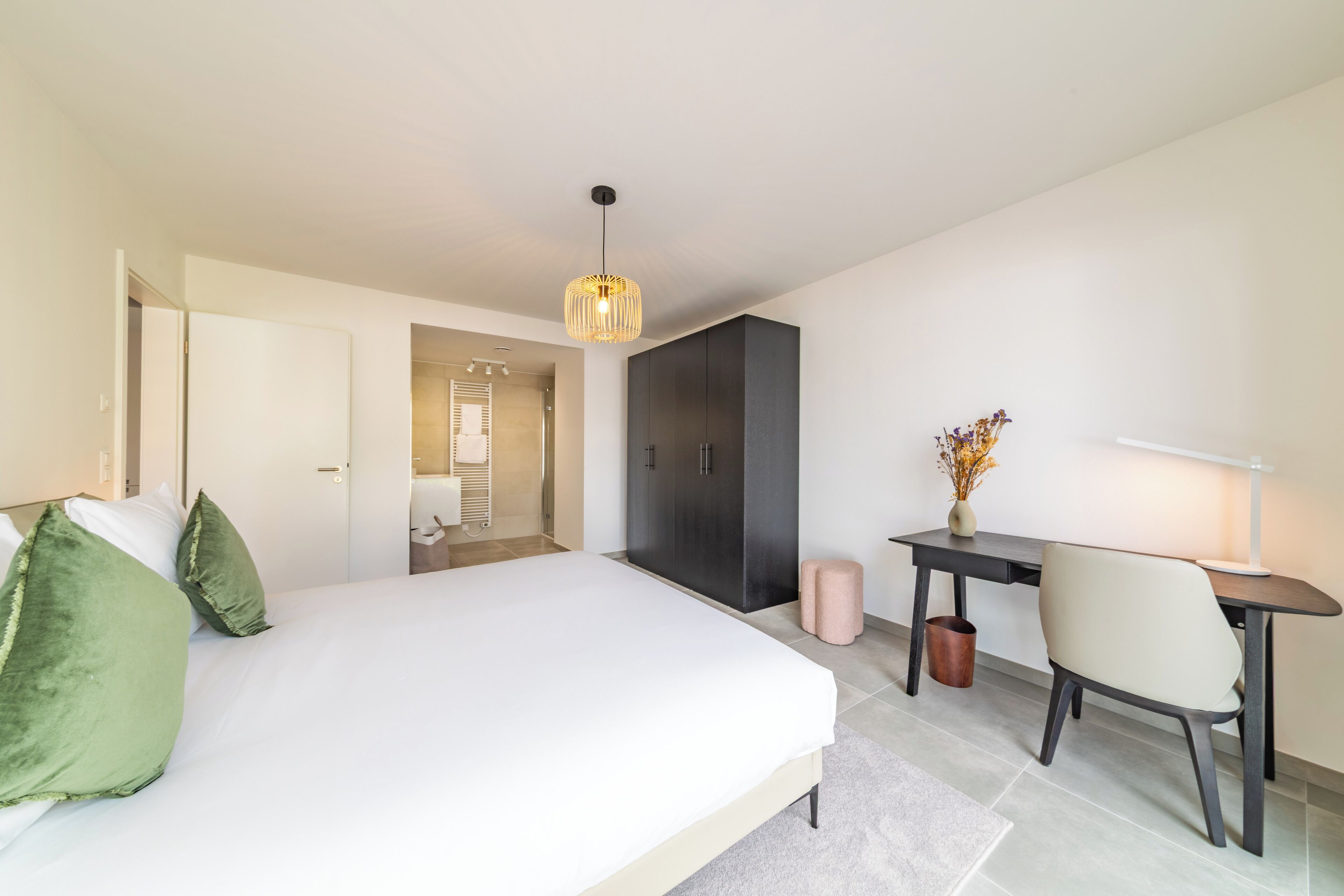 A modern hotel room with a large bed, a desk, and a chair. There is a door leading to a bathroom and a hallway.