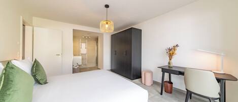 A modern hotel room with a large bed, a desk, and a chair. There is a door leading to a bathroom and a hallway.