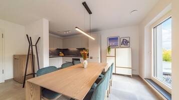 Zona pranzo moderna con tavolo in legno e sedie color petrolio, una cucina sullo sfondo e un'ampia finestra con vista.