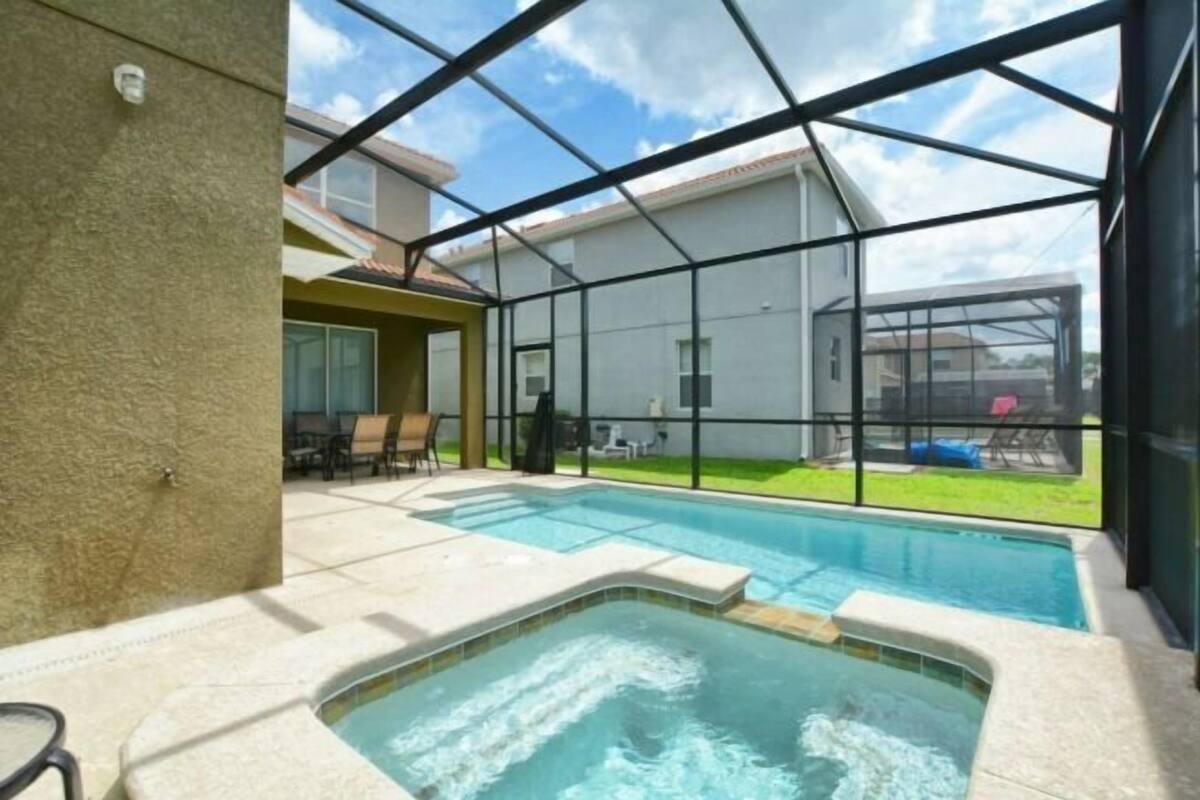 Piscina con jacuzzi, rodeada por un recinto de vidrio, con vista a una casa y jardín.
