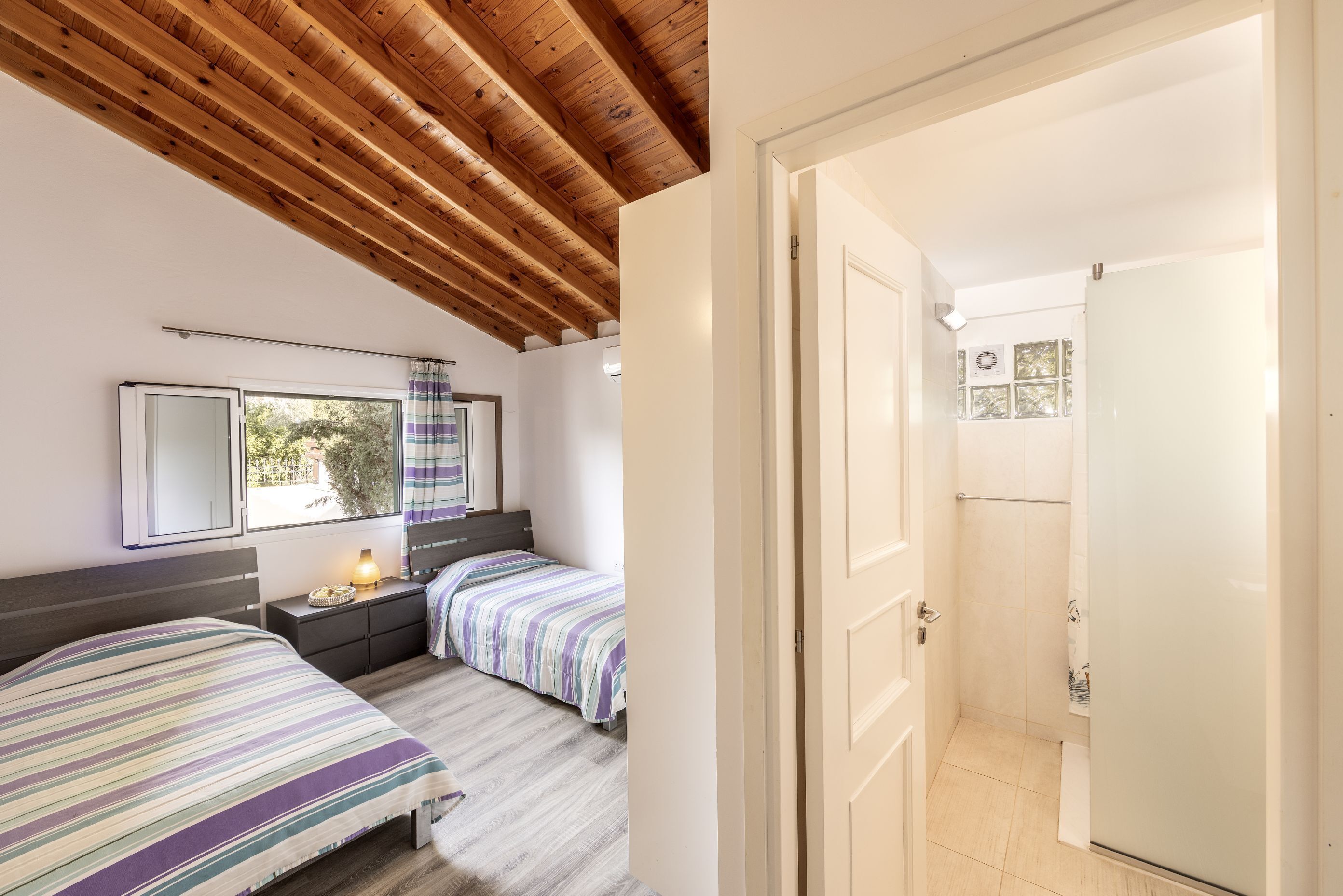 A bedroom with two beds, a wooden ceiling, a window with a view, and a bathroom visible through an open door.