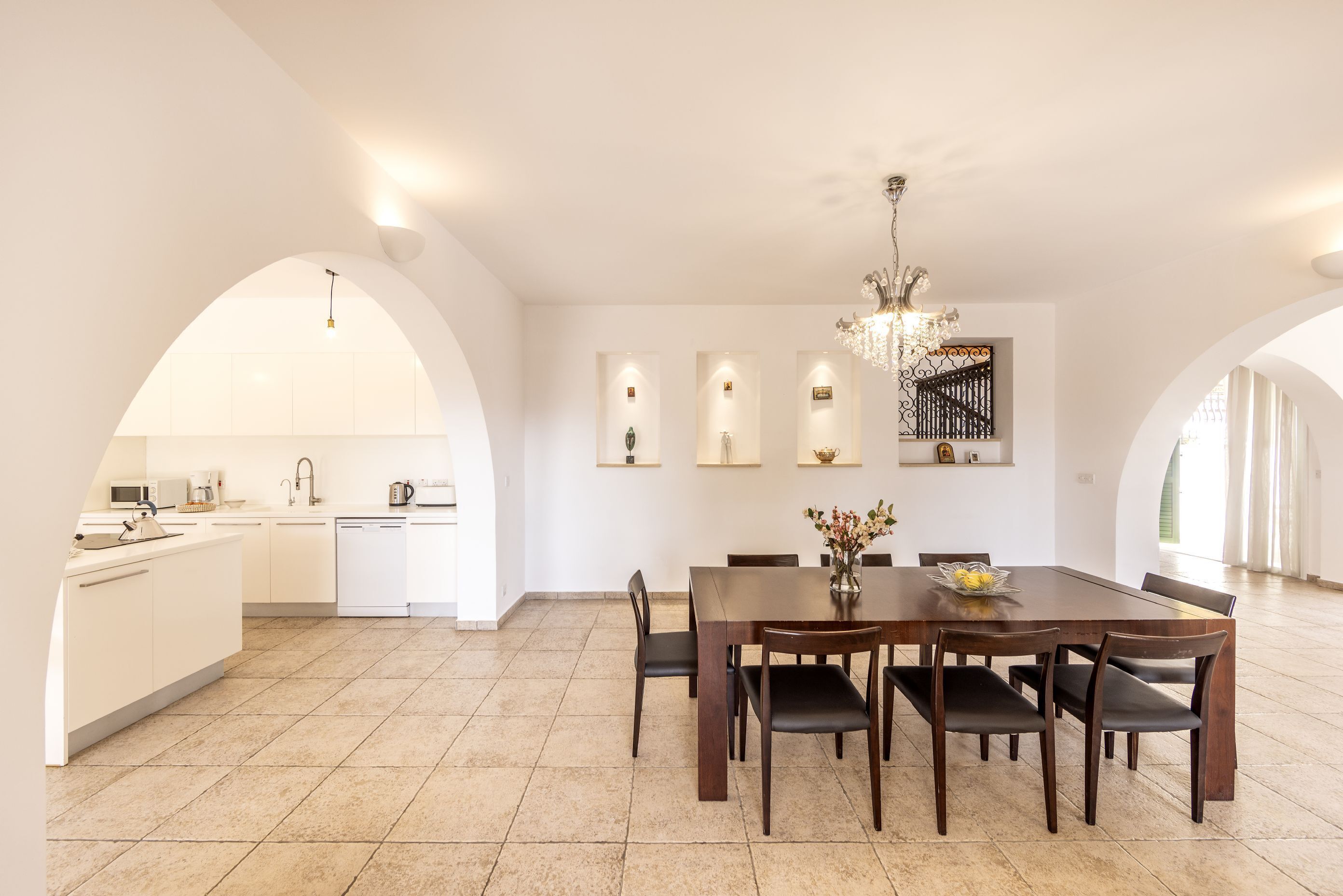 A spacious dining area with a large wooden table and chairs, a chandelier, and a kitchen in the background.