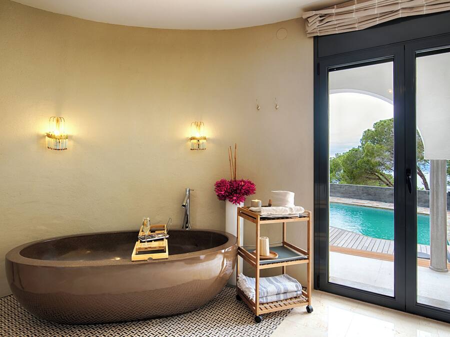 A modern bathroom with a freestanding bathtub, two wall sconces, a wooden shelf with toiletries, and a view of a pool through a glass door.