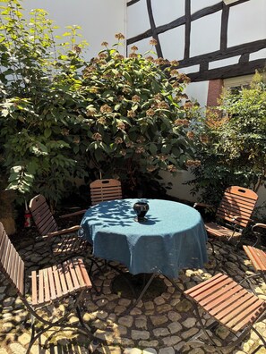 Een terras buiten met een ronde tafel, afgedekt met een blauwe tafelkleed, omringd door houten stoelen.