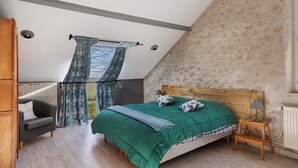 A bedroom with a sloped ceiling, a bed with a green bedspread, a wooden headboard, a nightstand with a lamp, a grey sofa, and a window with curtains.