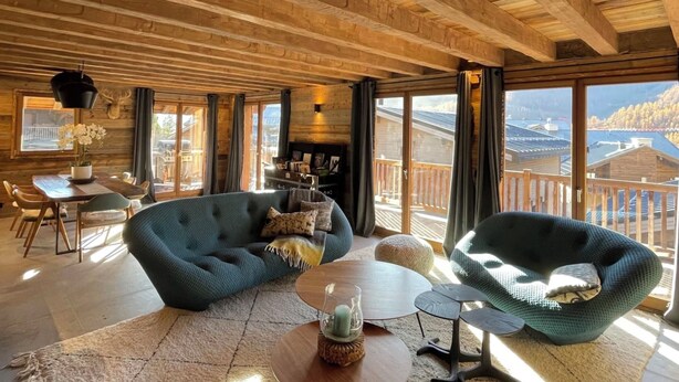 Una habitación amplia con un alto techo de madera, un sofá azul acogedor, una mesa de centro redonda y vistas a la montaña a través de grandes ventanales.