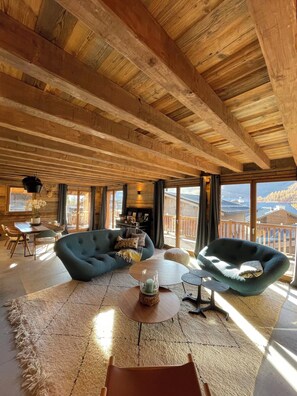 A spacious room with a high wooden ceiling, a cozy blue sofa, a round coffee table, and a view of the mountains through large windows.