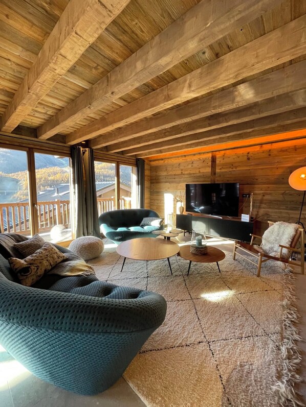 A cozy cabin interior with a wooden ceiling, a large sofa, a television, and a view of a mountainous landscape through large windows.
