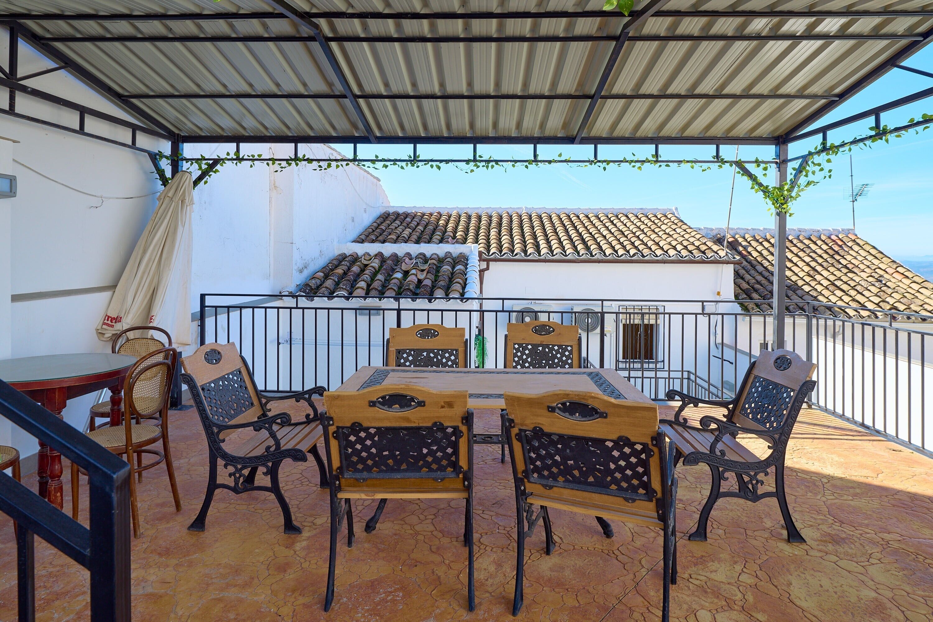 A covered patio with a table and chairs, a view of a building with a tiled roof, and a metal railing.