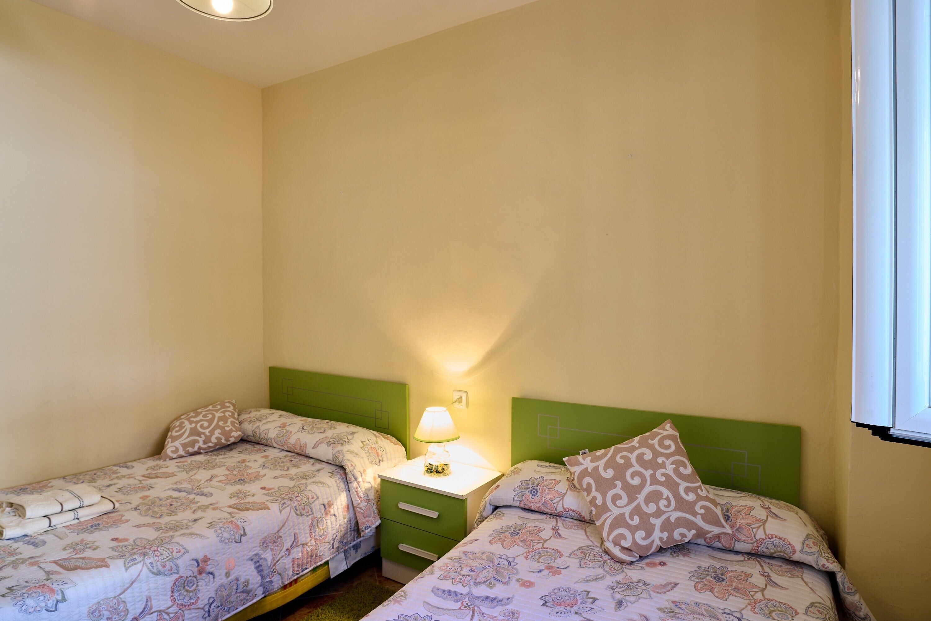 A bedroom with two beds, a nightstand with a lamp, and a window.