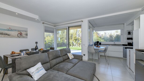 Ein modernes Wohnzimmer mit grauem Sofa, einem Essbereich mit Tisch und Stühlen sowie einer Küche mit weißen Schränken und einem Fenster.