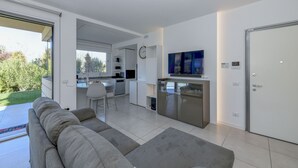 Ein modernes Wohnzimmer mit grauem Sofa, eine Essecke mit weißem Tisch und Stühlen, eine Küche mit Kühlschrank und Mikrowelle sowie ein großes Fenster mit Blick auf einen Garten.