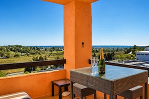Ein Balkon mit Korbmöbeln, einer Champagnerflasche und zwei Gläsern auf einem Tisch, mit Blick auf eine Küstenlandschaft.