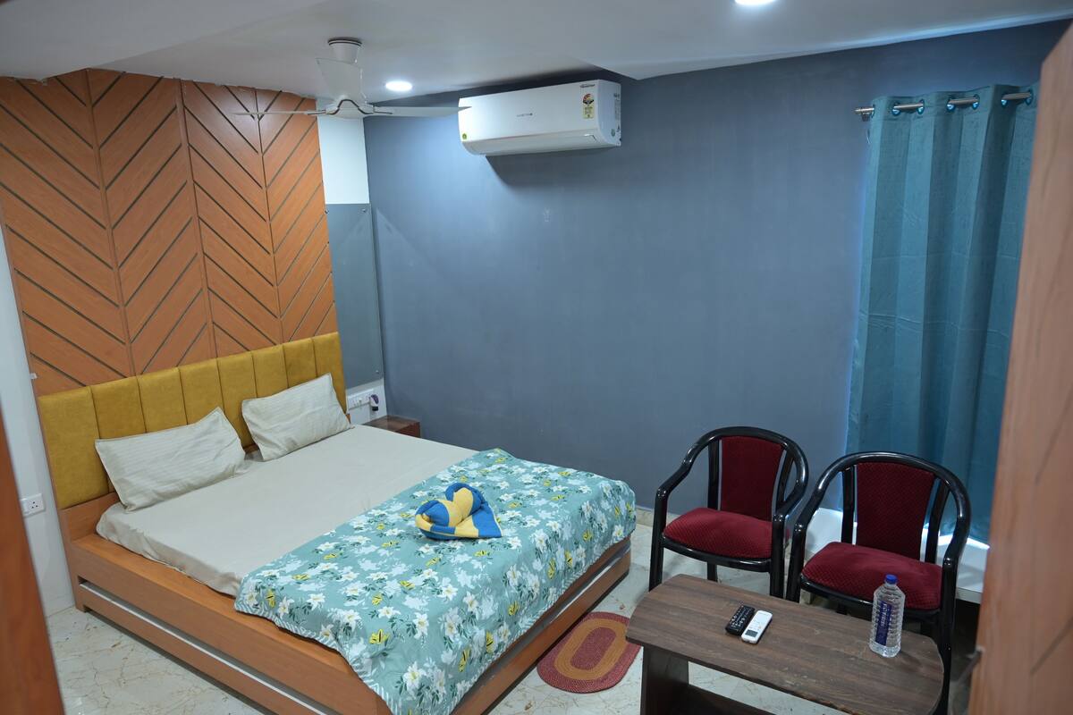 A hotel room with a bed, a desk, two chairs, an air conditioner, and a curtain.
