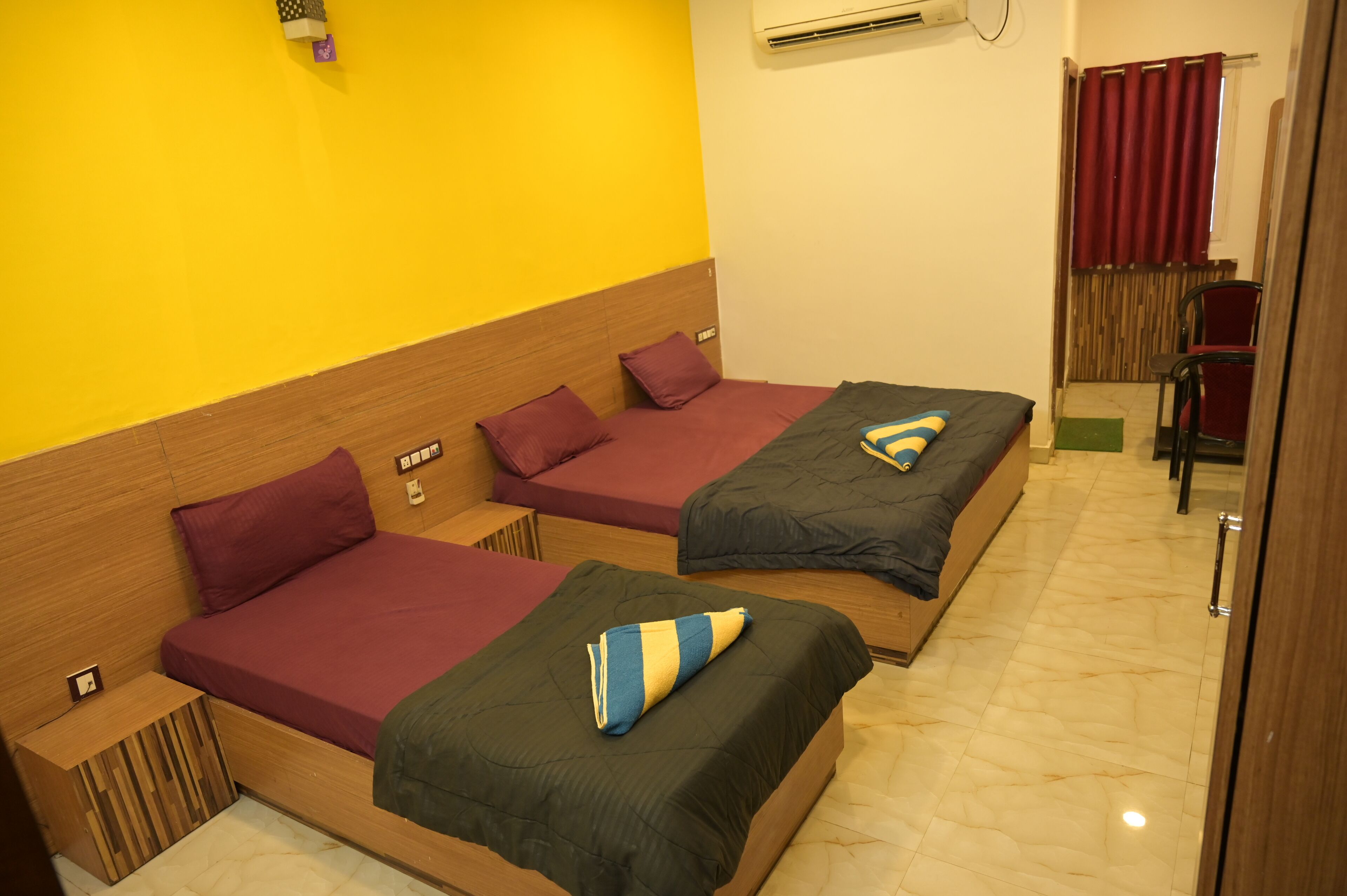 A hotel room with two beds, a wooden headboard, a red curtain, and a small table with a lamp.