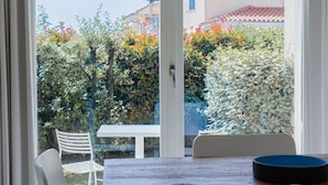 A view from a window with a table and chairs, a door, and a garden with various plants and a building in the background. - A welcoming space, ideal for two people (with the option of adding an infant), perfect for those seeking comfort and practicality. The bedroom offers a double bed—or comfortable single beds, depending on your needs—along with a small table and chairs (Porto San Paolo)