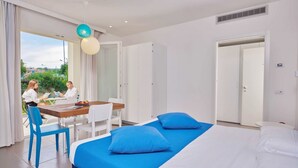 A hotel room with a large bed, two blue pillows, a wooden desk with chairs, and a view of greenery outside. - A welcoming space, ideal for two people (with the option of adding an infant), perfect for those seeking comfort and practicality. The bedroom offers a double bed—or comfortable single beds, depending on your needs—along with a small table and chairs (Porto San Paolo)