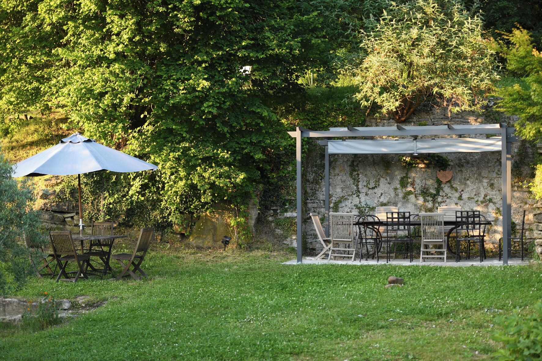 Eine Terrass mit Tisch und Stühlen, eine Pergola mit weißem Baldachin und eine Steinwand mit Grünpflanzen.