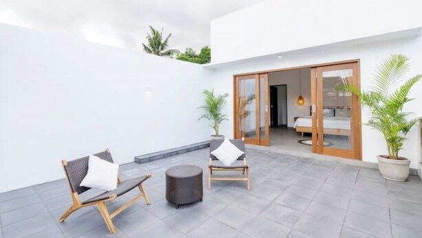 Un espacio moderno al aire libre con dos sillas y un reposapiés redondo, una puerta de madera que da a un dormitorio y plantas en macetas.