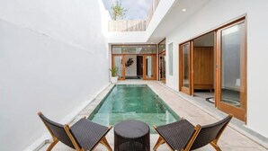 Un área de piscina moderna al aire libre con dos sillas de mimbre y una pequeña mesa redonda, rodeada por un muro blanco y una terraza de madera.