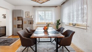 A dining area with a wooden table, four chairs, a fruit bowl, and a window with sheer curtains. - Alpine View Apartment with Mountain Views, Balcony & Wi-Fi (Schopfheim)