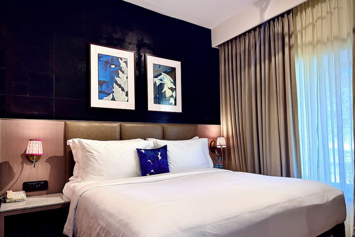 Ein Hotelzimmer mit einem großen Bett, zwei gerahmten Kunstwerken an der Wand, einem Nachttisch mit einer Lampe und einem Fenster mit Vorhängen.