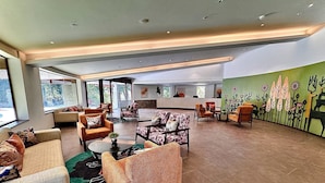 A spacious lobby with multiple seating areas, a reception desk, and large windows offering views of the outdoors. - The Corbett Hideaway - By Leisure Hotels (Ramnagar)