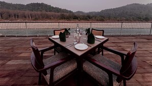 A dining table set for four with chairs, positioned on a wooden deck overlooking a river and hills. - The Corbett Hideaway - By Leisure Hotels (Ramnagar)